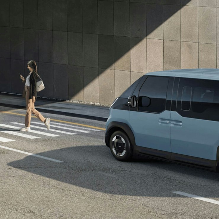 Kia PV5 WAV auf einer Stadtstrasse mit aktivem Frontkollisionswarner, der dabei hilft, einen Fussgänger voraus zu erkennen.