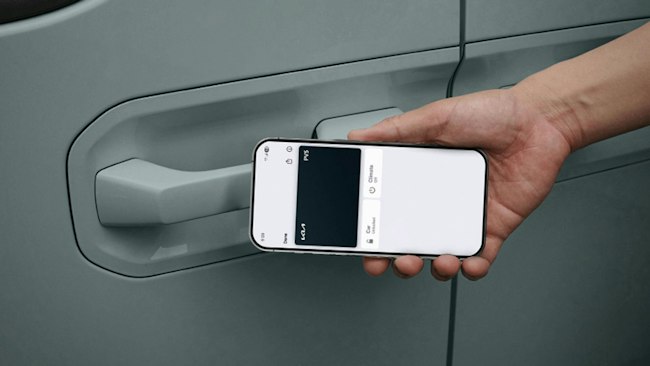 Eine Hand hält ein Smartphone mit dem Digital Key des Kia PV5 vor dem Fahrzeug.