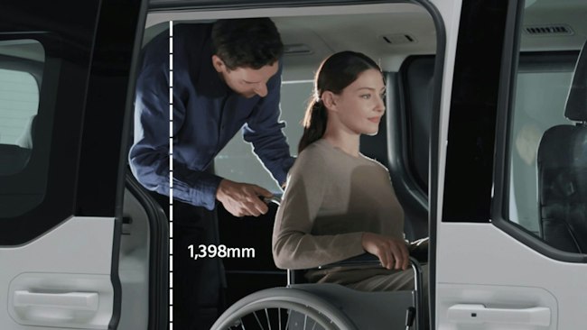 Eine Person im Rollstuhl fährt durch die Seitentür in den Kia&nbsp;PV5&nbsp;WAV und wird dabei von einer Person im Fahrzeug unterstützt.